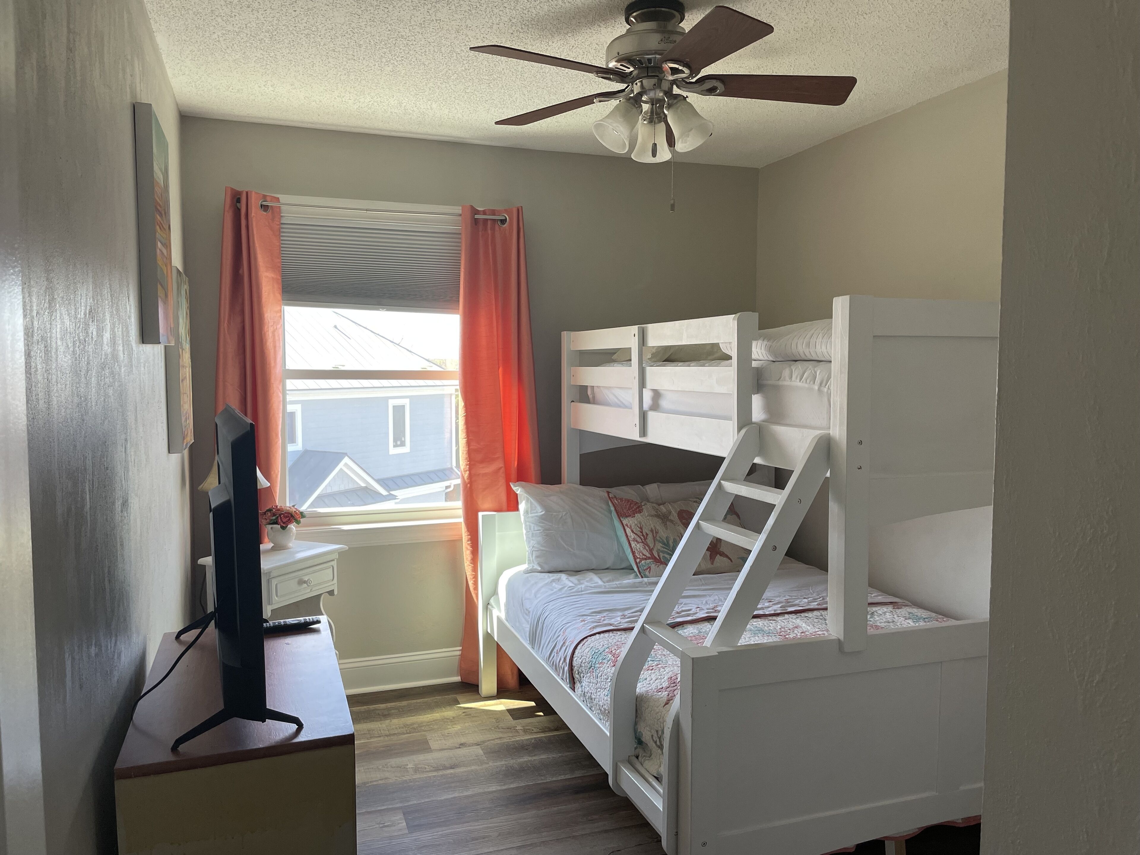 Un dormitorio con literas, un escritorio con ordenador, un ventilador de techo y una ventana con cortinas.