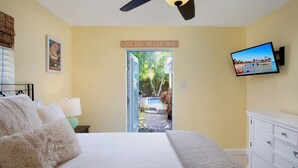 A bedroom with a bed, a TV mounted on the wall, a sign reading "SANDY TOES AND SALTY KISSES", and a view of a swimming pool and outdoor area. - BAREFOOT BUNGALOW~2 Bedroom, 2 Bathroom Home w/ Office, Pool and Tiki Hut! (Key West)