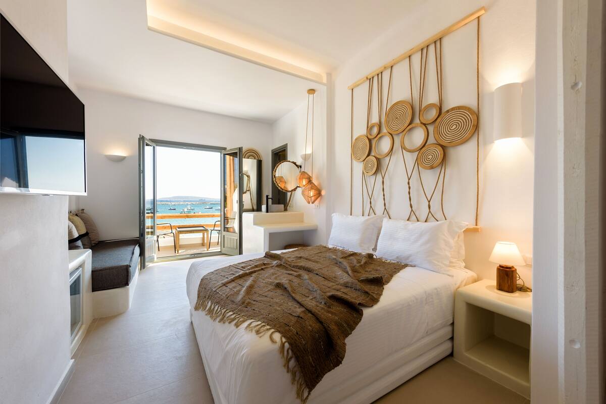 A modern bedroom with a large bed, a bedside table with a lamp, a wall-mounted decorative piece, and a view of the outdoors.