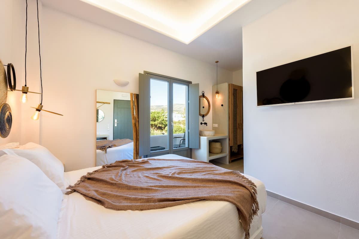 A bedroom with a bed, a large window, a mirror, a television, and a bathroom with a sink and a towel rack.