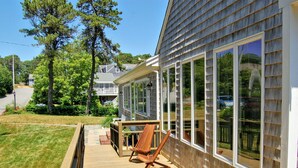 A wooden deck with a chair and a table, overlooking a backyard with a lawn and trees. - Spacious, private cottage just a few minutes walk to Ridgevale Beach! (Chatham)