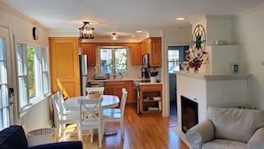 A modern kitchen with wooden cabinets, a dining table with chairs, a fireplace, and a view of the outdoors through large windows. - Spacious, private cottage just a few minutes walk to Ridgevale Beach! (Chatham)