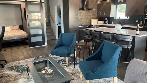 Un salon moderne comprenant un canapé, deux fauteuils bleus, une table basse et un coin cuisine avec un bar et des tabourets.