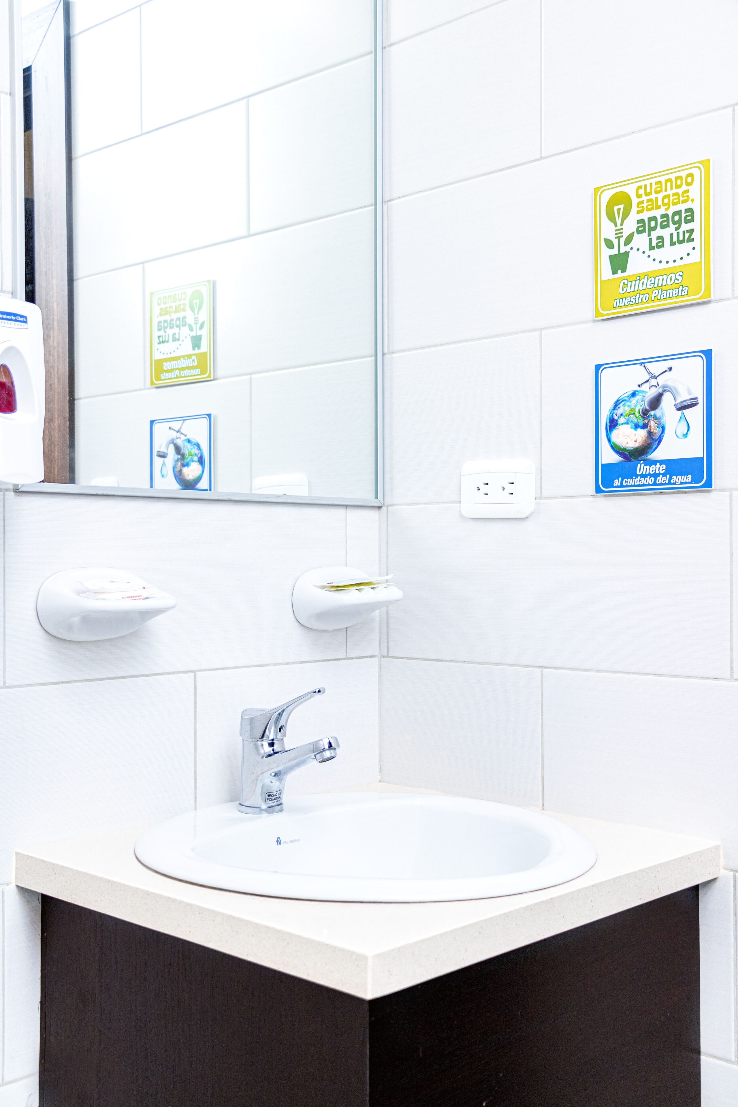 A bathroom with a sink, mirror, and two soap dispensers. There are two signs on the wall promoting water conservation.
