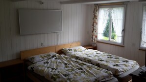 Ett rum med två enkelsängar, en sänggavel i trä, ett litet nattduksbord, ett fönster med blommiga gardiner och en väggmonterad ljusströmbrytare.