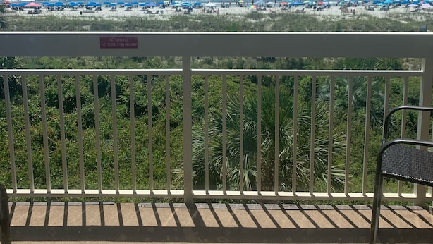 Uma varanda com vista para a praia, um aviso proibindo churrascos e toalhas no corrimão, e uma cadeira.