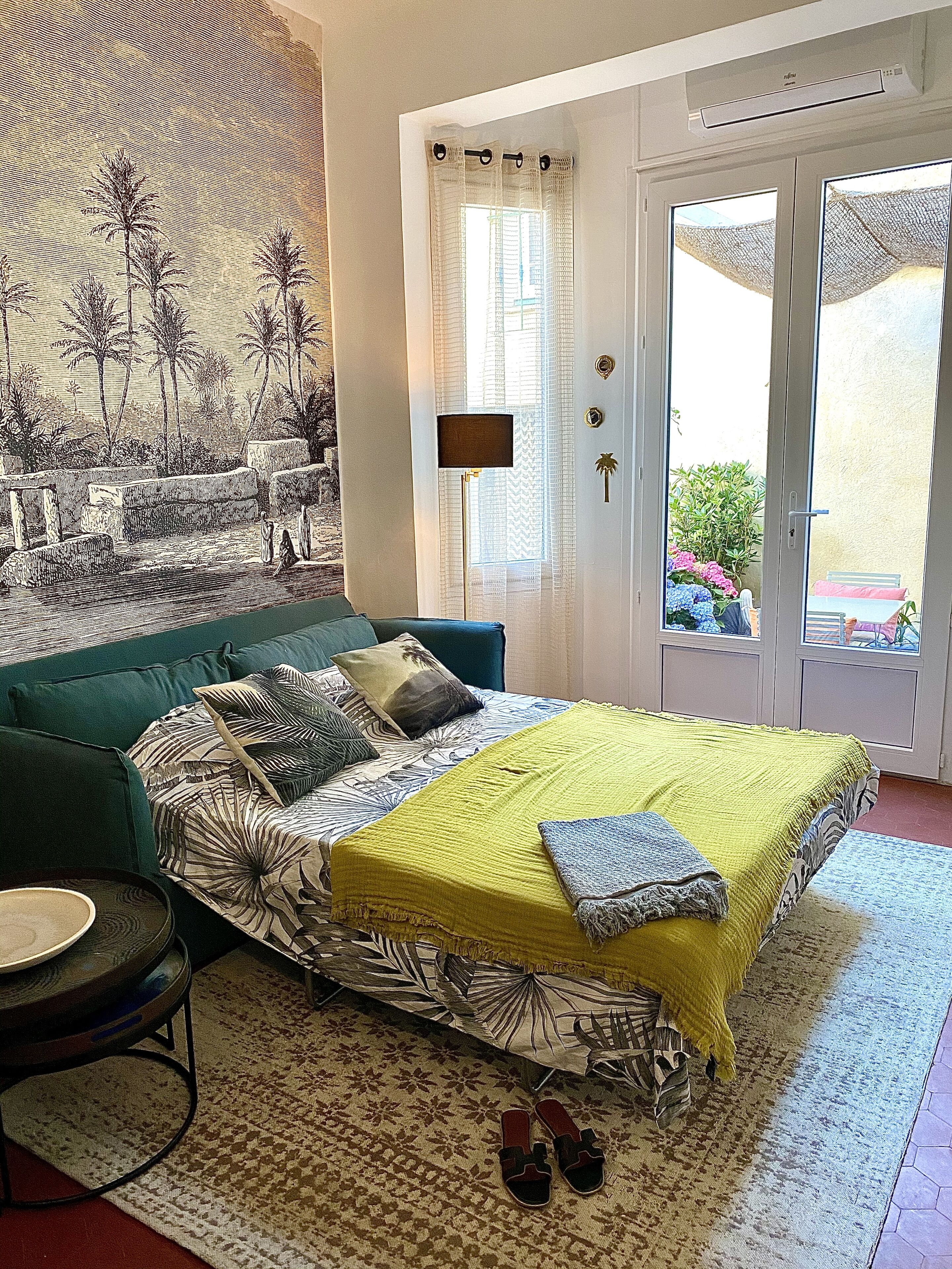 Un dormitori amb un llit, un sofà verd, una taula petita i una paret amb un mural tropical.