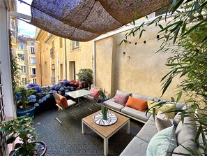 An outdoor seating area with a table, chairs, and a sofa. There are plants and flowers around, and a building is visible in the background. - Dar Saad, a hanging garden in the historic center of Aix (Aix-en-Provence)