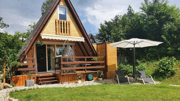 Una pequeña casa de madera en forma de triángulo con un porche, una hamaca colgante y una sombrilla de patio.