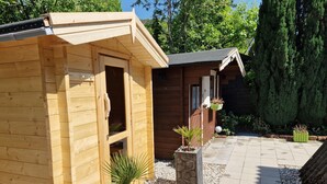 Una cabaña de madera con un pequeño porche, una jardinera de piedra con una planta de palma, y un camino que lleva a una terraza de madera.