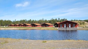 Une rangée de cabanes rouges, avec un petit quai s’avançant dans un lac, le tout entouré d’une forêt.