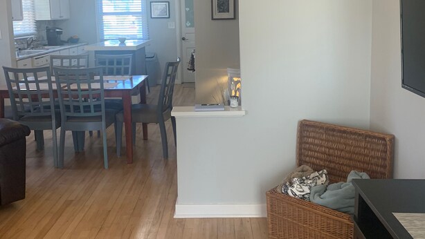 Uma sala de estar ampla com piso de madeira, uma mesa de jantar com cadeiras, um pequeno aparador branco, um banquinho de vime e uma televisão sobre um suporte.