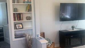 Uma sala de estar com uma estante branca, uma TV de tela plana, um rack de entretenimento preto e piso de madeira.