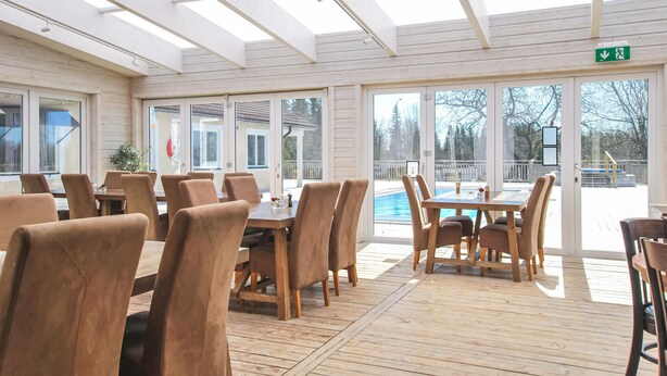 A sunlit indoor dining area with wooden tables and chairs, large windows, and a view of an outdoor swimming pool. - 1 bedroom nice home in Kosta (Kosta)