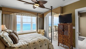 Een hotelkamer met een bed, een televisie op een houten kast, een balkon met uitzicht op de oceaan en een plafondventilator.