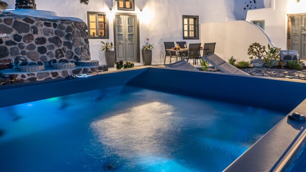 A swimming pool with stone structures and a dining area with chairs and tables. - Noelia Suites (Santorini)