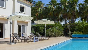 Zona junto a la piscina con sillones de descanso y sombrillas, una piscina azul cristalina y un edificio con tejado de baldosas y ventanas.