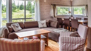 A cozy cabin interior with a wooden ceiling, large windows offering a view of a forest, a sofa, a striped chair, and a dining table. - Stunning home in Sjusjøen with sauna (Sjusjøen)