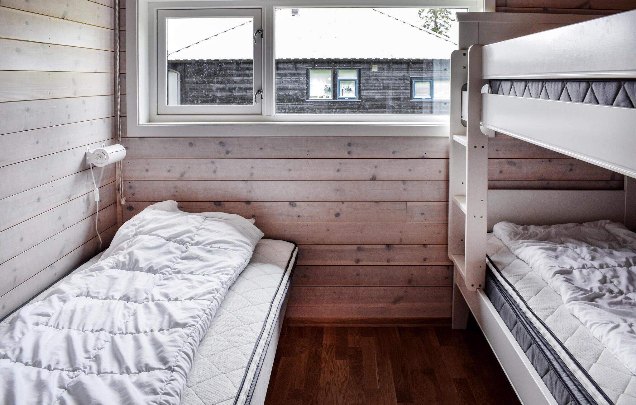A small, compact room with two bunk beds, wooden walls, a window with a view of a building, and a wooden floor.