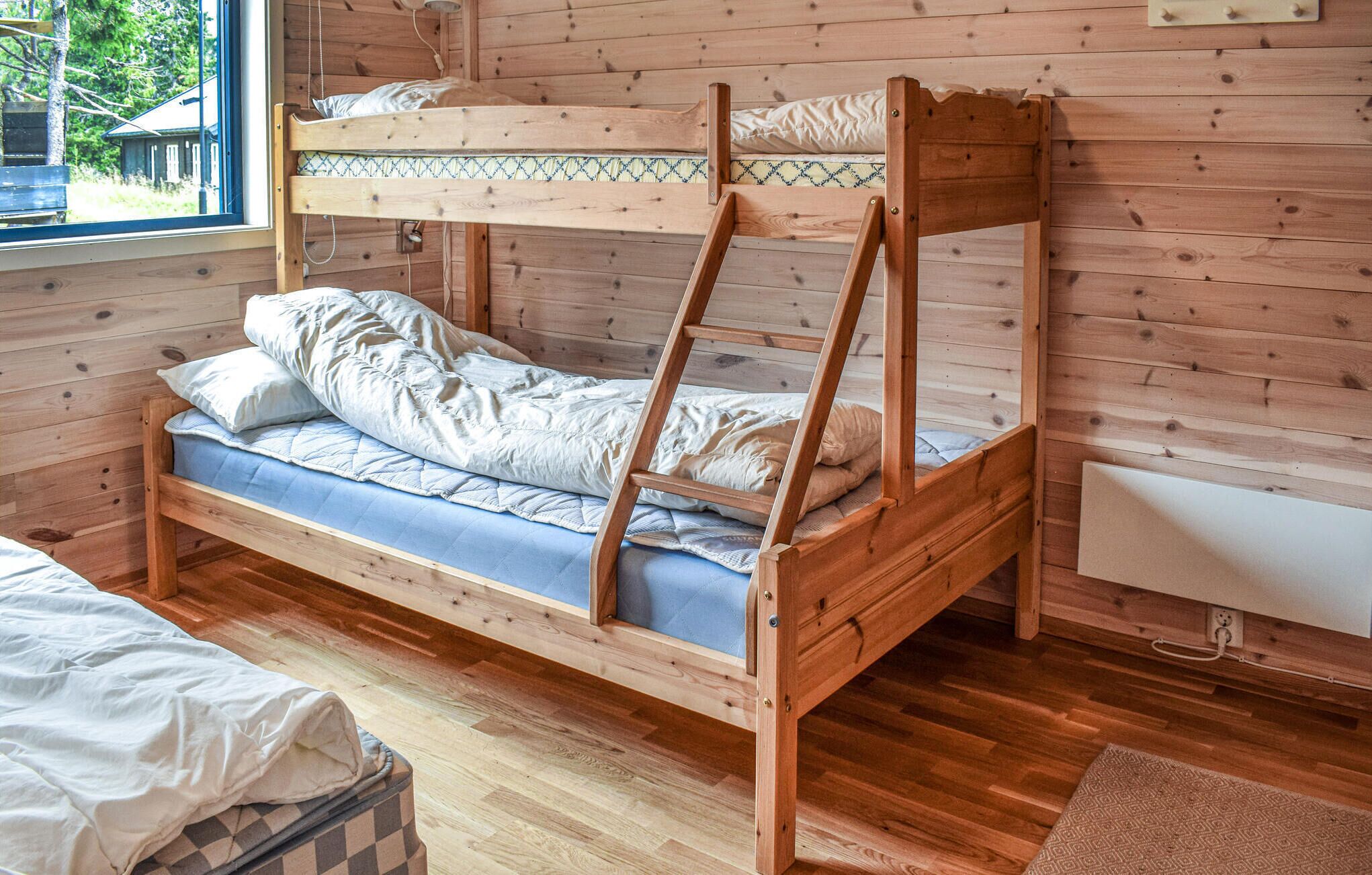 A bunk bed with a ladder, a single bed, and a small bedside table in a wooden room with a window.