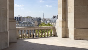 Vista desde un balcón con barandillas de piedra, que da a un paisaje urbano con edificios y la Torre Eiffel a lo lejos.