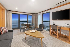 A modern hotel room with a large window, a flat-screen TV, a desk, a sofa, and a dining table.