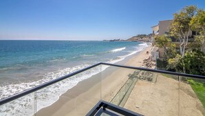 Vista a la playa desde un balcón con barandilla, cielo despejado y una costa distante.