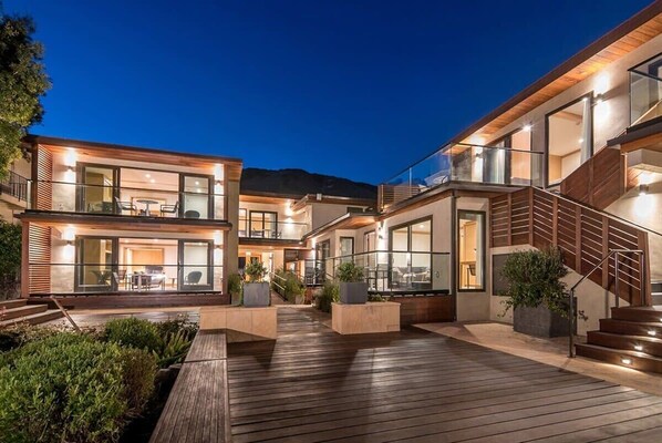 Une maison moderne à plusieurs étages, dotée d’une terrasse en bois, d’un espace salon extérieur et de grandes fenêtres.