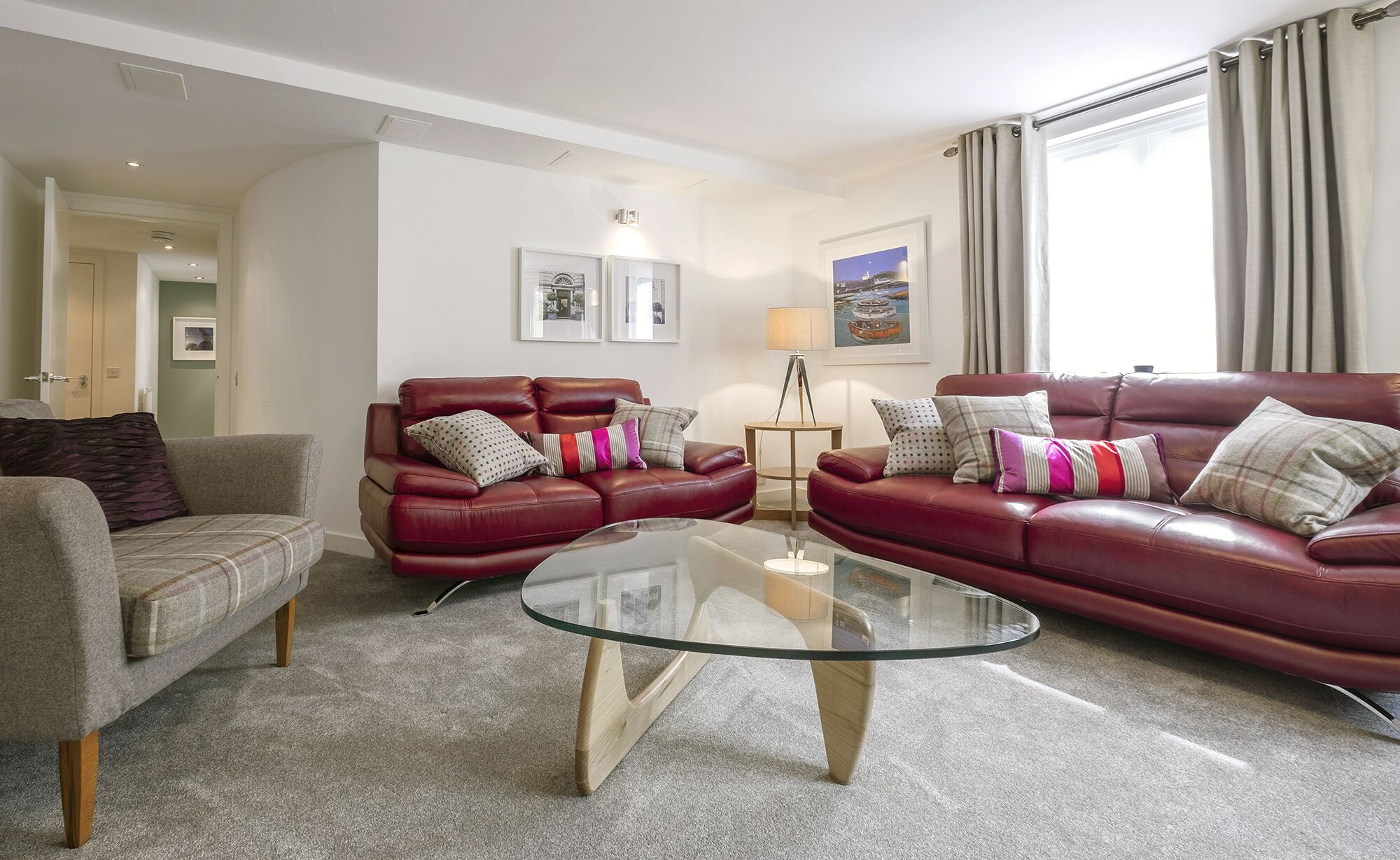 Un salon avec un ensemble de canapés en cuir rouge, une table basse en verre et un fauteuil gris. Deux tableaux encadrés ornent le mur, une lampe est posée sur une table d’appoint, et une fenêtre avec des rideaux offre une vue sur l’extérieur.