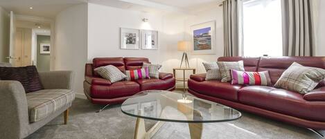 Un salon avec un ensemble de canapés en cuir rouge, une table basse en verre et un fauteuil gris. Deux tableaux encadrés ornent le mur, une lampe est posée sur une table d’appoint, et une fenêtre avec des rideaux offre une vue sur l’extérieur.