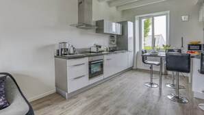 A modern kitchen with white cabinetry, stainless steel appliances, and a built-in oven. There are two bar stools and a window providing natural light. - Kerelen - Maison familiale avec jardin proche plage - Carantec (Carantec)