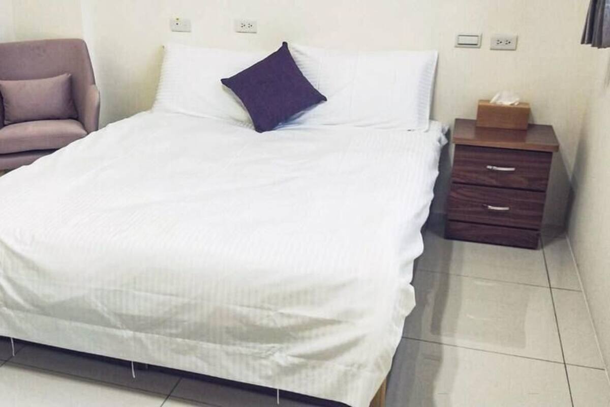 A neatly made bed with a white comforter and a dark blue pillow, a small wooden nightstand with a tissue box, and a purple armchair.