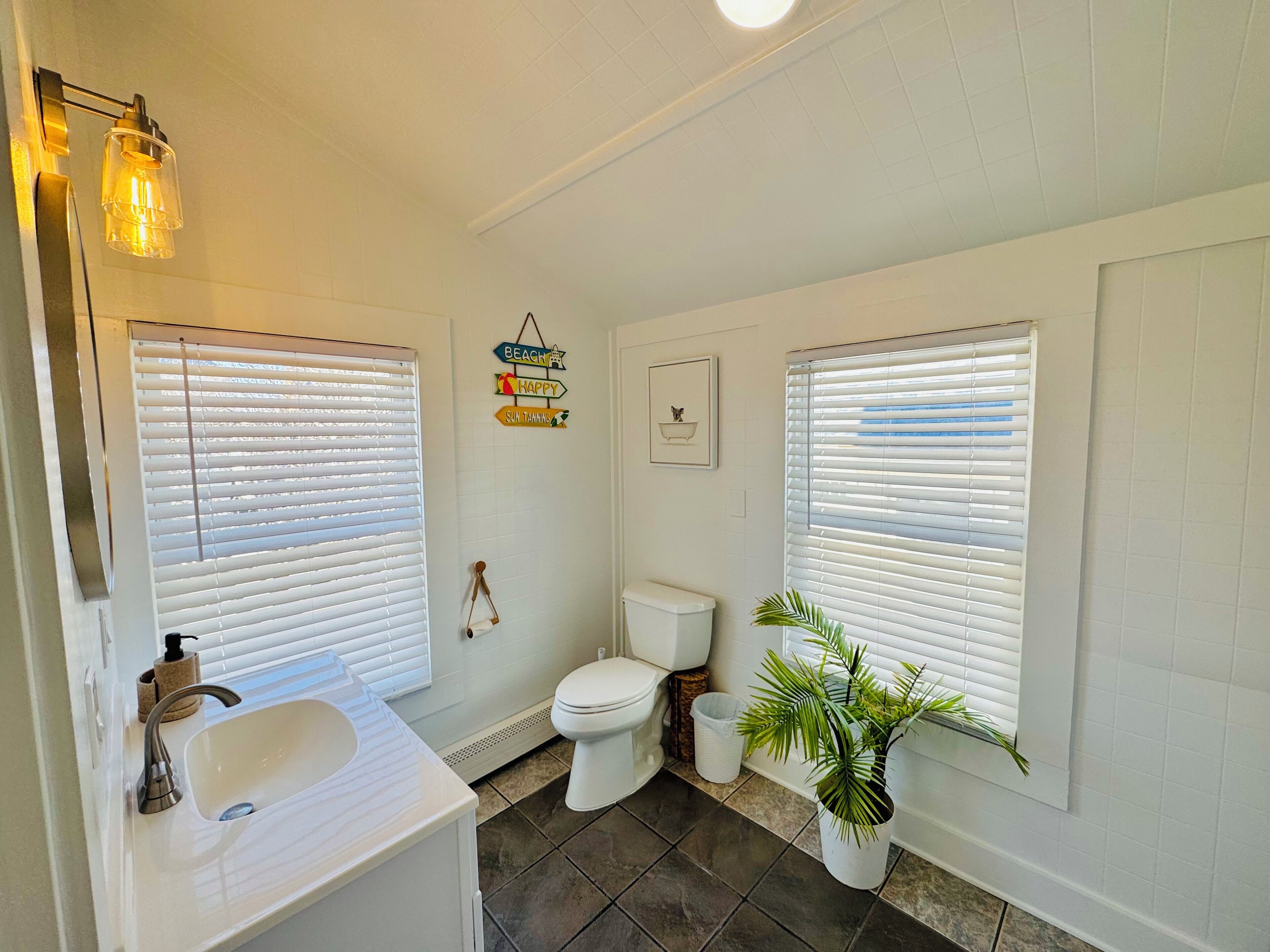 A bathroom with two sinks, a toilet, and a shower. There are decorative signs on the wall and a potted plant.