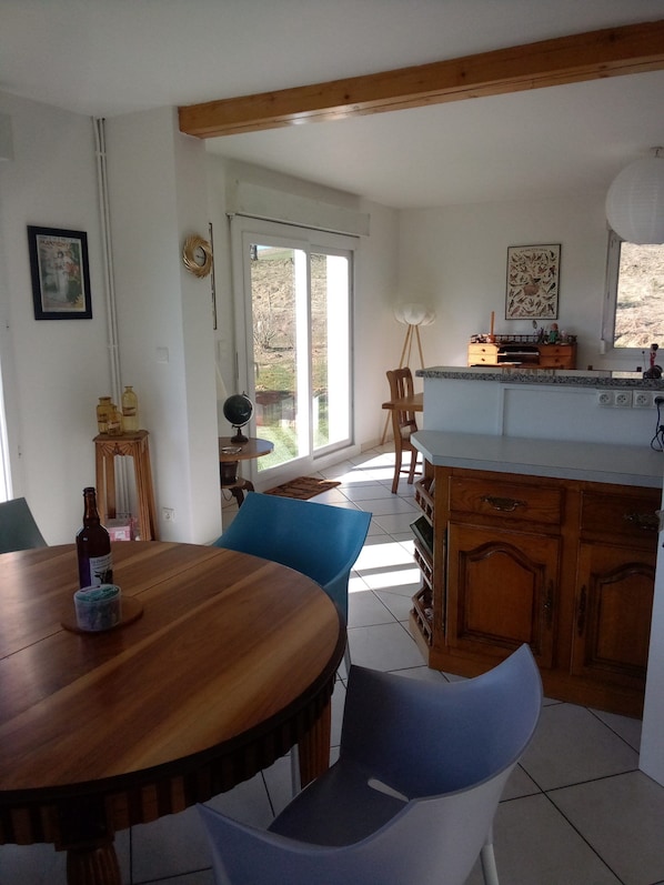 Un coin repas avec une table et des chaises en bois, une cuisine avec des meubles en bois et, vue à travers une porte vitrée, un espace extérieur.