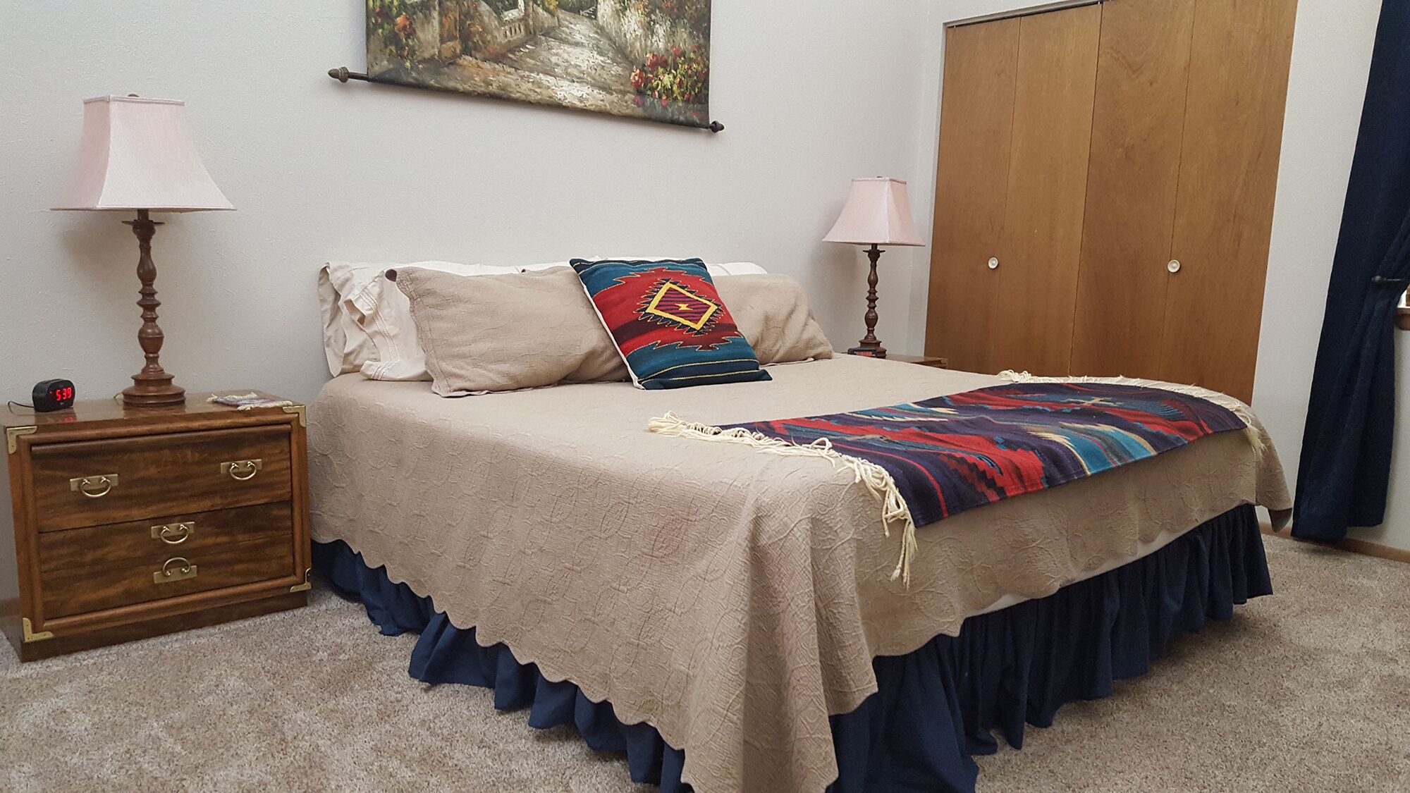 Une chambre à coucher avec un lit, deux lampes de chevet, une commode en bois, un tableau au mur et une armoire.