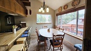 A dining area with a wooden table, chairs, and a chandelier. There is a window with a view of trees and a building outside. - Close to the Lifts, Sleeps Up to 8, Windows and Deck with Ski Mountain Views (Angel Fire)