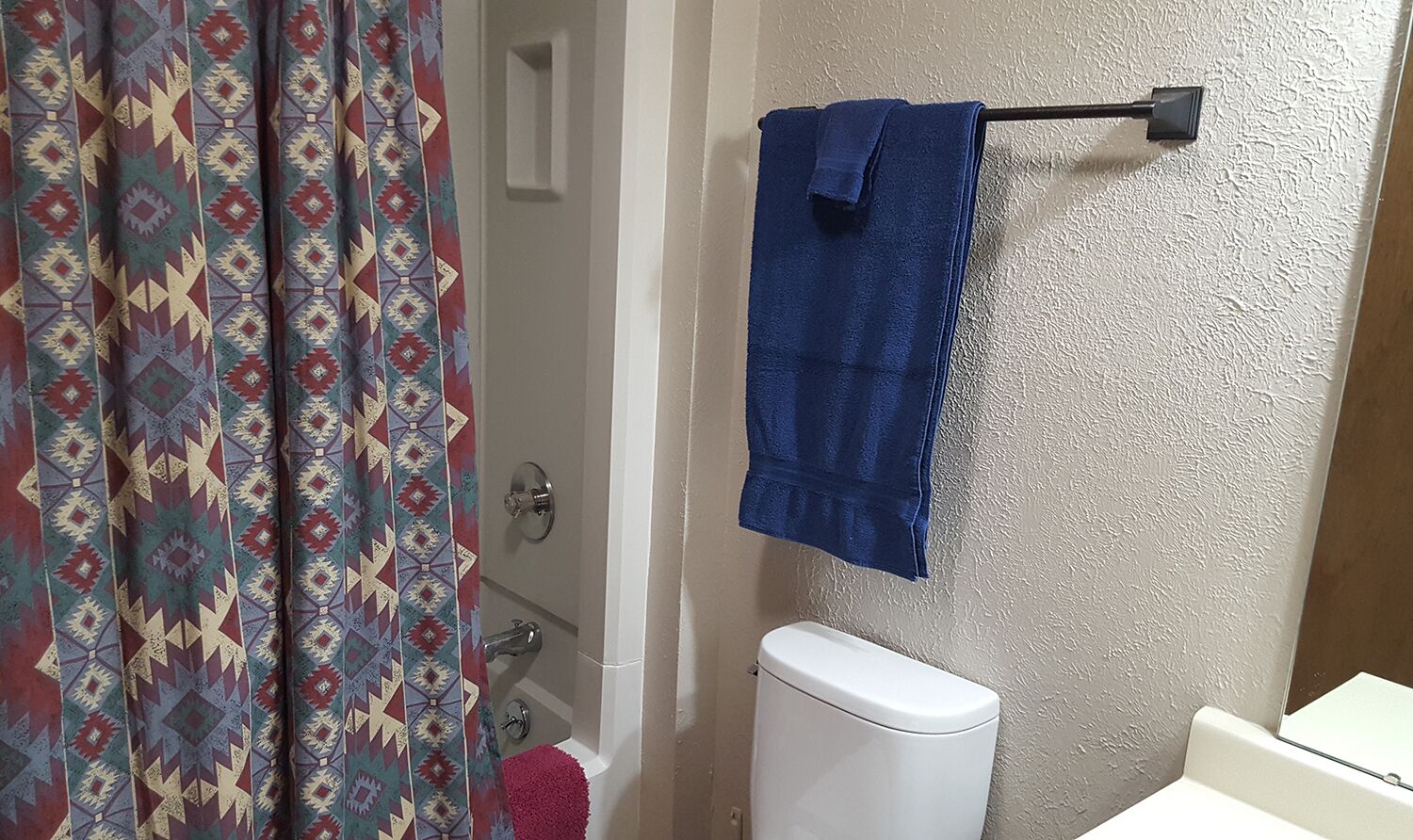 Et badeværelse med et hvidt toilet, et blåt håndklæde hængende på en håndklæderadiator, og en bruseforhæng med et geometrisk mønster.