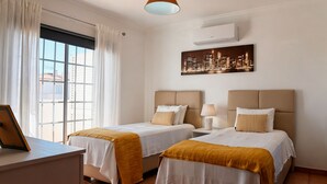 Ein Hotelzimmer mit zwei Betten, einem Schreibtisch, einem Stuhl, einem Fenster mit Vorhängen, einer Klimaanlage und einem Stadtbild an der Wand.