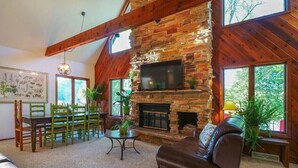 A cozy living room with a stone fireplace, wooden beams, a television, and a dining area with green chairs. - Walk to Road America Races. Enjoy the lake frontage and fishing and kayaking. (Plymouth)