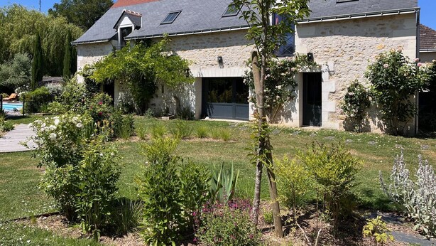 Uma casa de pedra com telhado inclinado, uma pequena garagem anexa e um jardim bem cuidado com diversas plantas e flores.