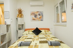 Une chambre d’hôtel bien rangée, avec un lit, une fenêtre, un climatiseur fixé au mur et une étagère sur laquelle se trouvent un vase et un document encadré.