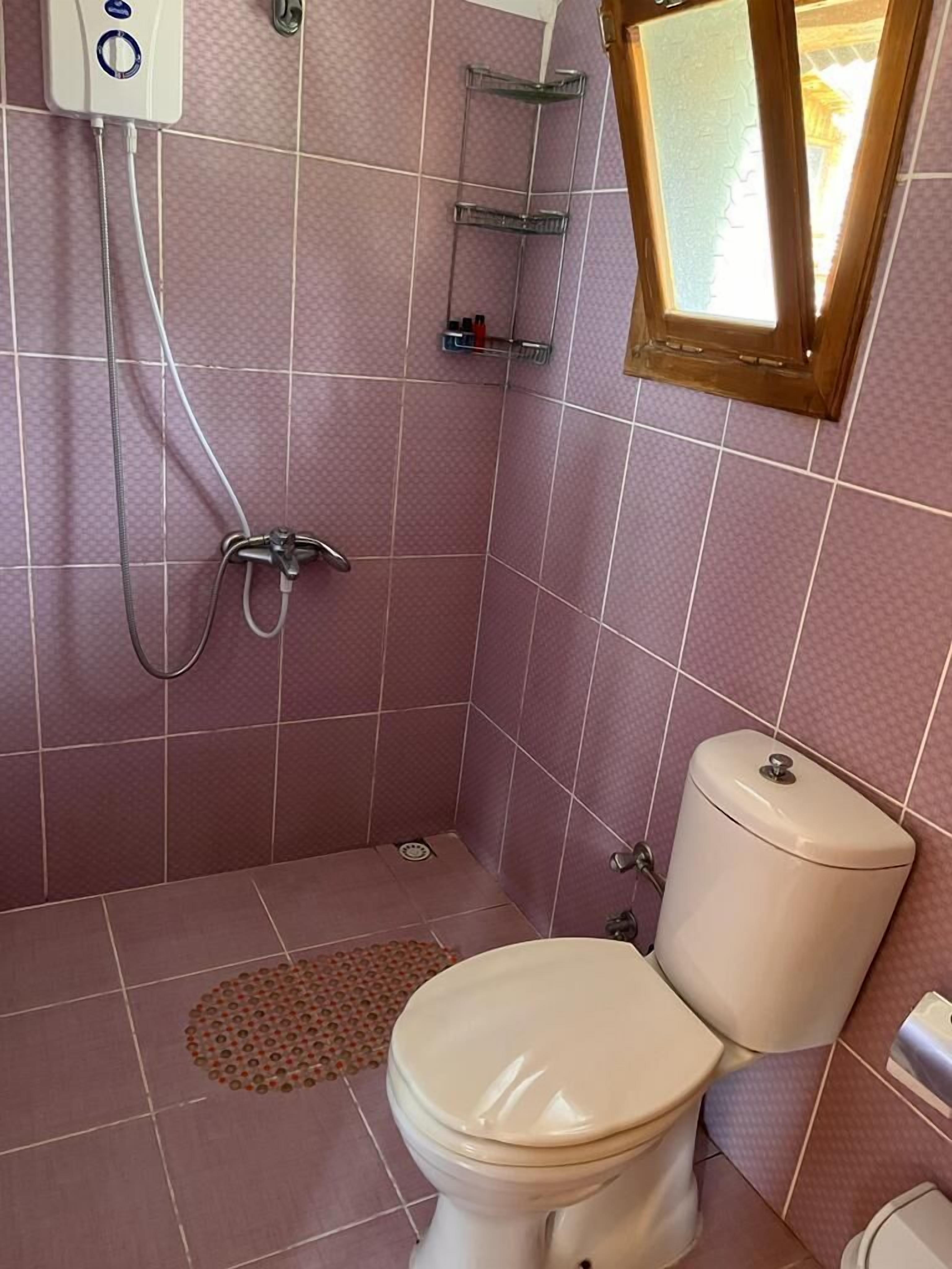 Une salle de bain avec des murs recouverts de carreaux roses, des toilettes, une douche et une fenêtre.