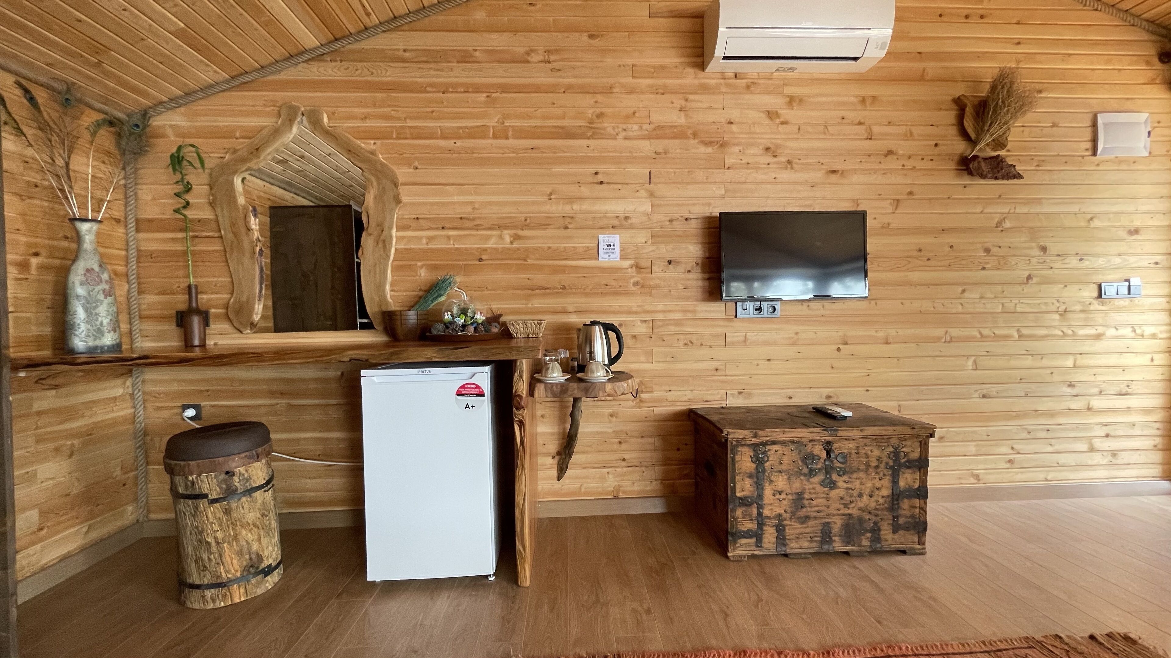 A wooden cabin interior with a refrigerator, a television mounted on the wall, a rustic wooden chest, and a small table with a kettle.