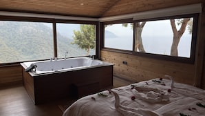Una habitación con una bañera de madera grande, una cama con cubrecama blanco y vistas a un paisaje montañoso a través de amplios ventanales.