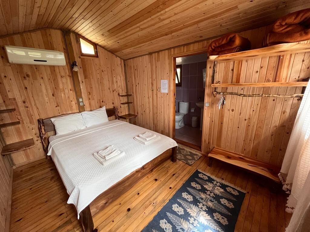 Quarto de cabana de madeira com uma cama, guarda-roupa de madeira e um banheiro visível através de uma porta aberta.