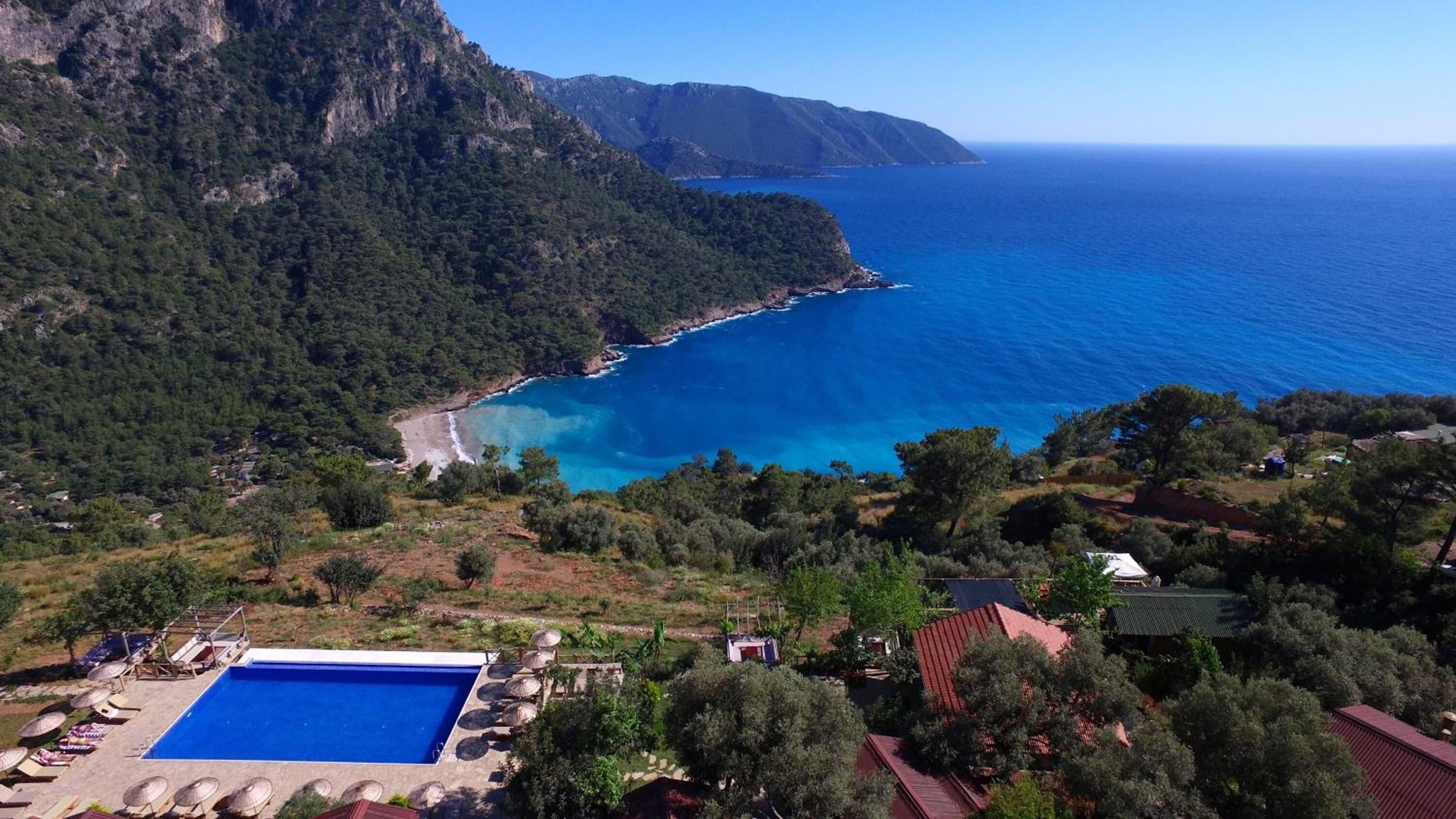 Un resort costero con piscina, acceso a la playa y montañas al fondo.