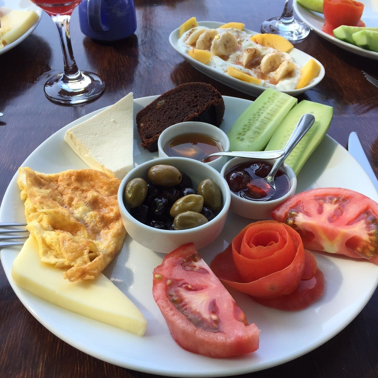A plate with various foods including toast, a slice of cake, olives, tomatoes, and a bowl of jam.