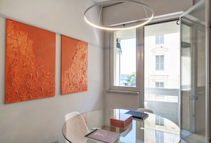 Une pièce moderne avec une table en verre, deux tableaux abstraits, un luminaire au plafond et une vue à travers une porte vitrée.