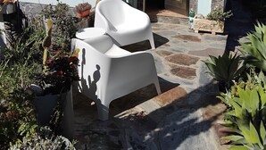 Un bâtiment avec une façade en pierre, un toit recouvert de tuiles et une porte. On y voit des plantes en pot et une chaise en plastique blanche à l’extérieur.