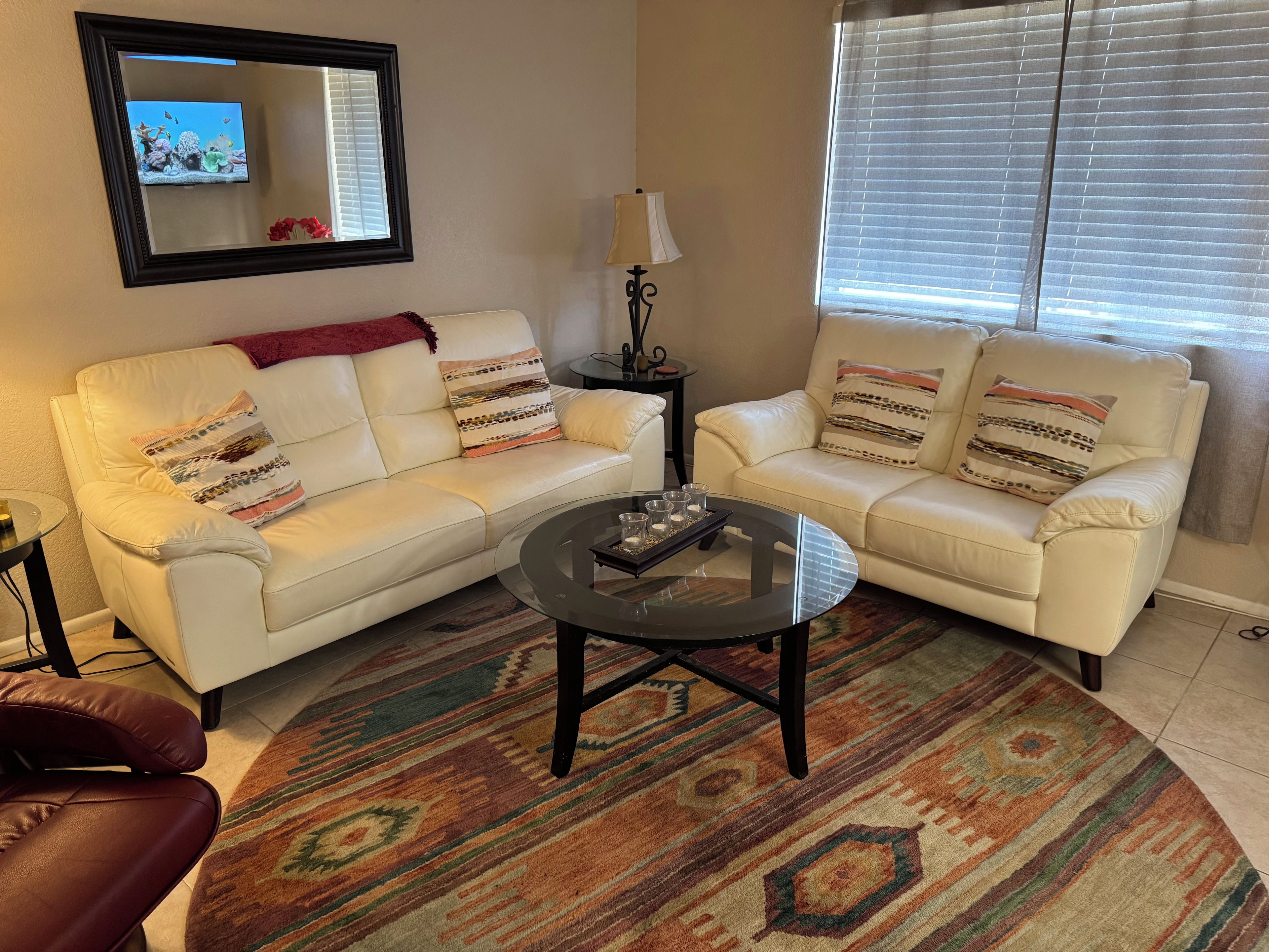 A living room with two white leather sofas, a glass coffee table, a patterned rug, a lamp, and a television on the wall.
